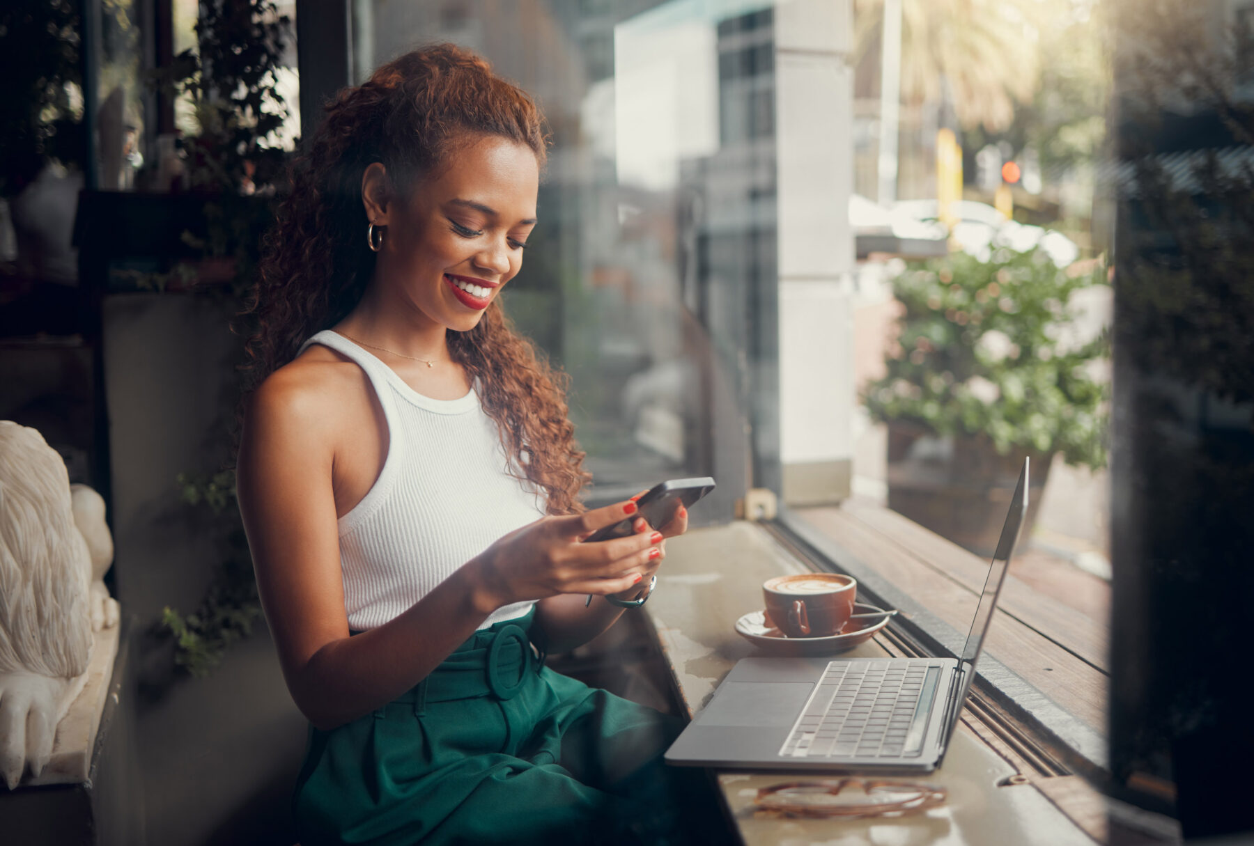 A woman sits by a window in a cafe, smiling at her smartphone. A laptop and a cup of coffee are on the table in front of her. Sunlight filters in, and greenery can be seen outside.